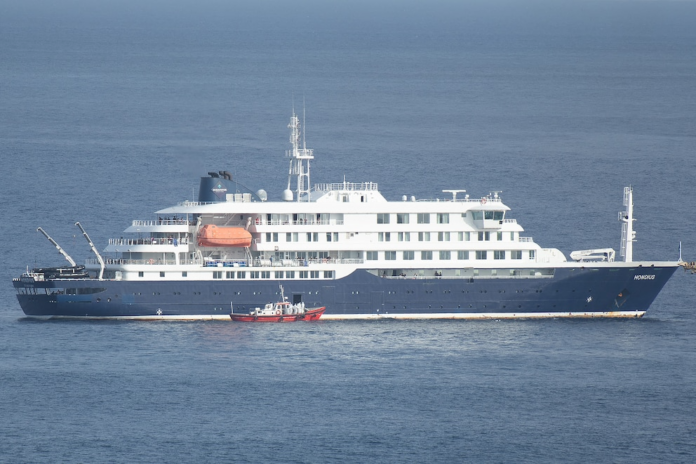 Una lancha ambulancia con tripulantes vestidos con trajes de protección se aproxima a la puerta de práctico en el costado de estribor del crucero MV Hondius, mientras permanece detenido frente al puerto de Praia, la capital de Cabo Verde, el 5 de mayo de 2026, mientras los pasajeros observan desde los puentes. (AFP)